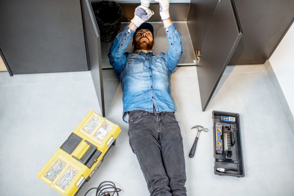 Handyman repairing kitchen plumbing lying under the sink on the floor, view from above